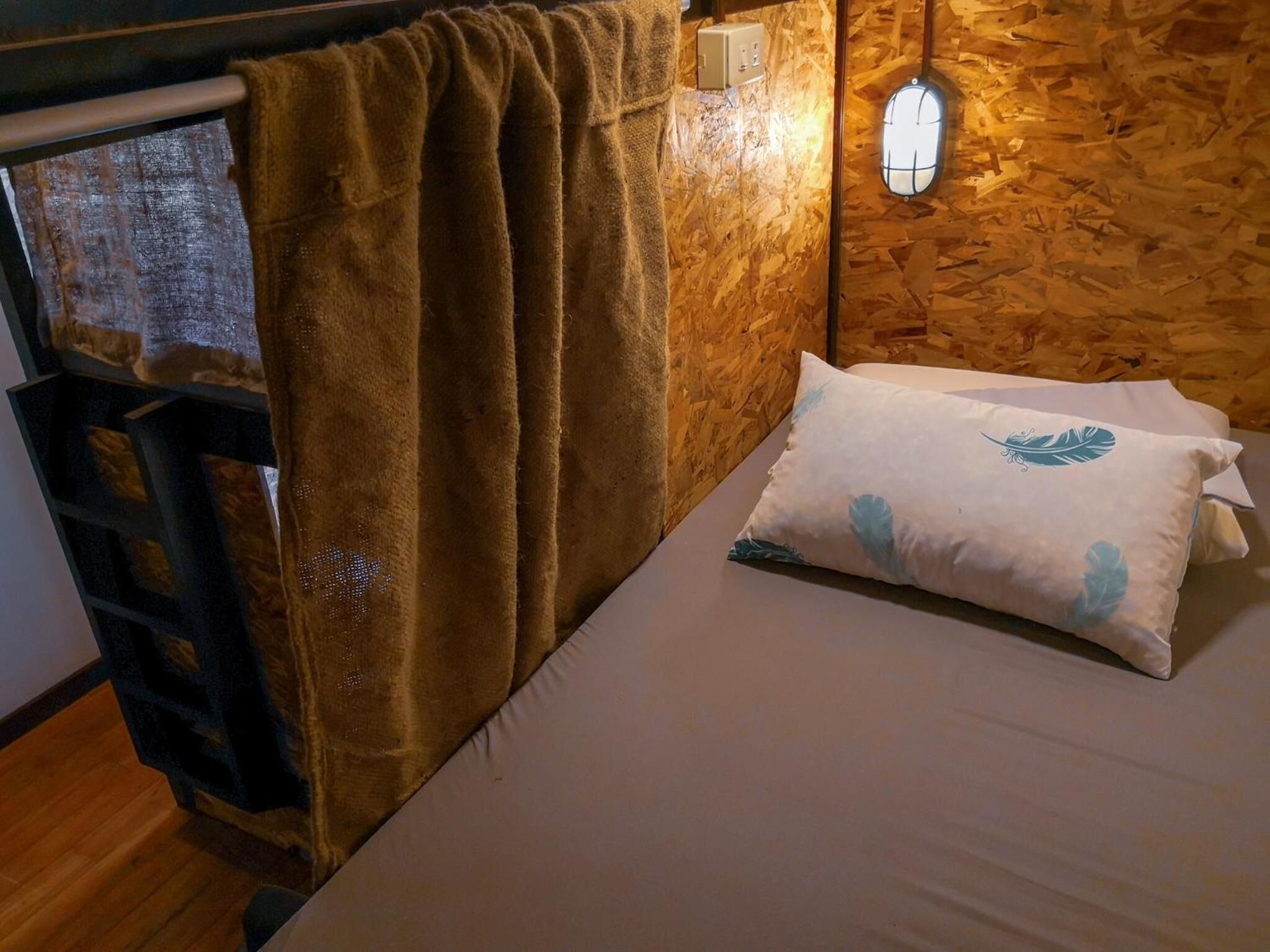 Una cama con un almohadón blanco y una cortina marrón en una habitación con paredes y suelo de madera.