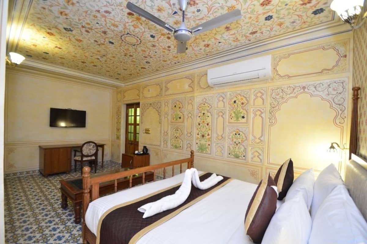 A hotel room with a bed, a desk, a chair, a ceiling fan, and a TV.