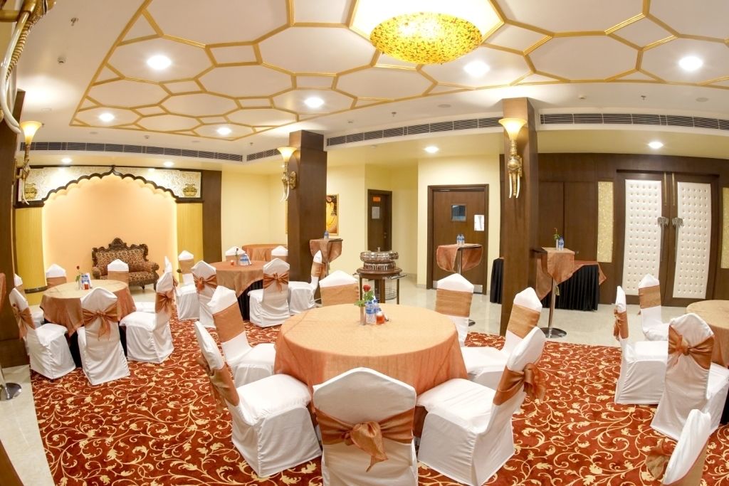 Uma sala de conferências com mesas redondas, cadeiras brancas com fitas marrons e um tapete vermelho estampado.
