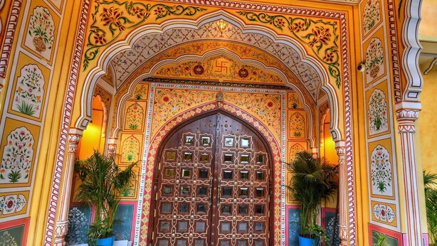 An ornate entrance with a large wooden door, intricate floral and geometric patterns on the walls and ceiling, and potted plants on either side. - Nirbana Palace - A Heritage Hotel (Jaipur)
