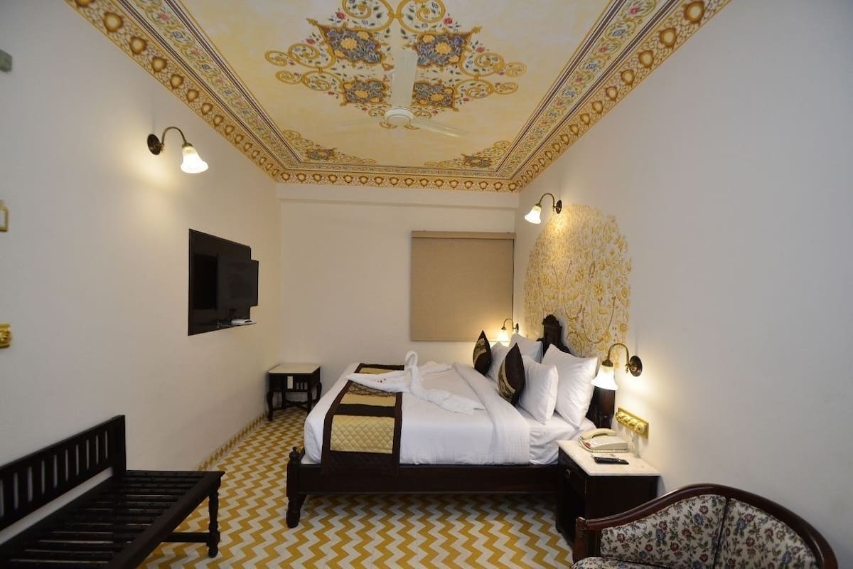 A hotel room with a bed, a bedside table with a lamp, a chair, a small table, and a decorative ceiling.