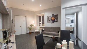 A modern living room with a grey sofa, a dining table with candles, a potted plant, and a large window with blinds. - Rental In Rome Apartment Archimede (Rome)
