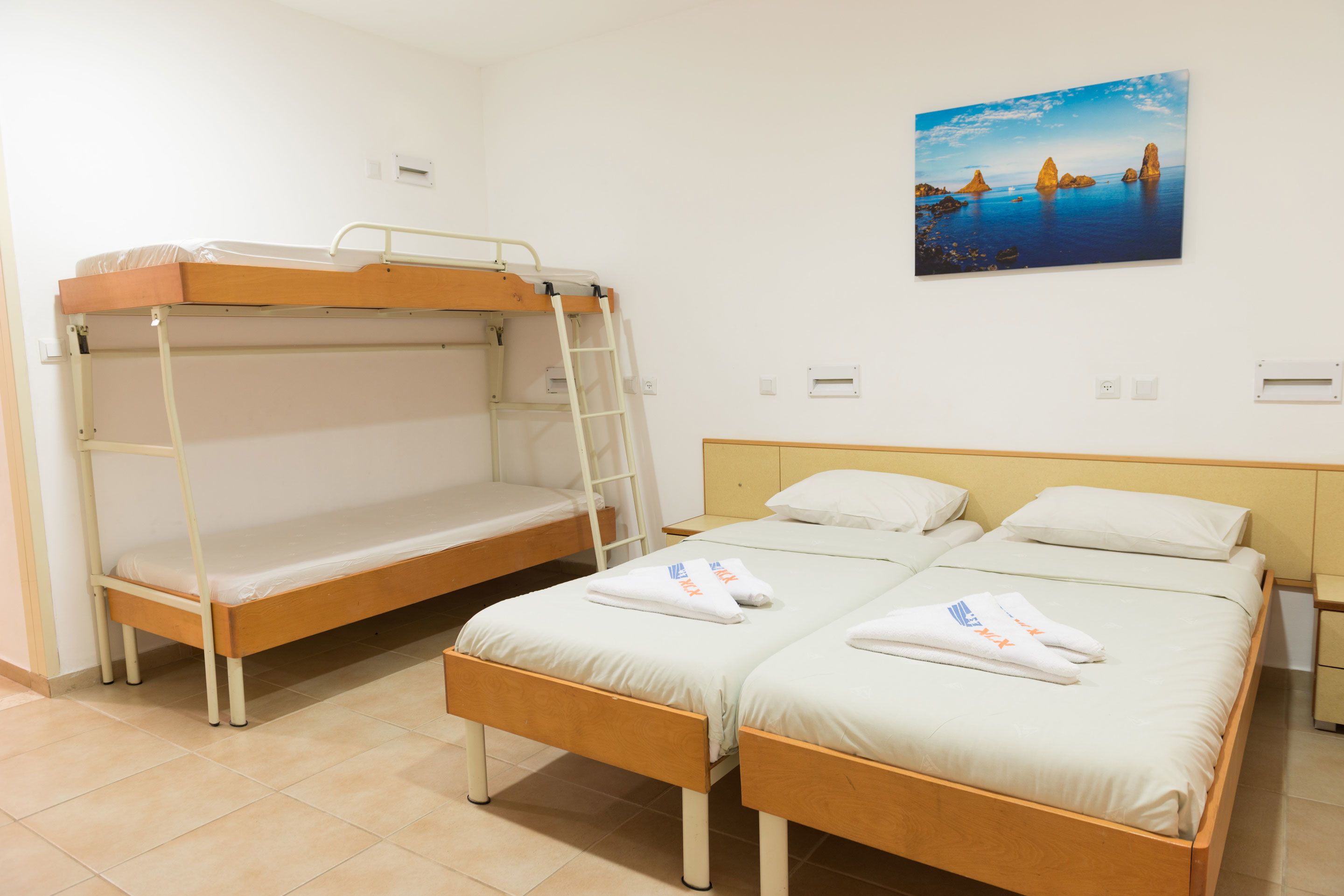 Una habitación con dos camas individuales y una litera, un cuadro de un paisaje costero y suelo de baldosas.