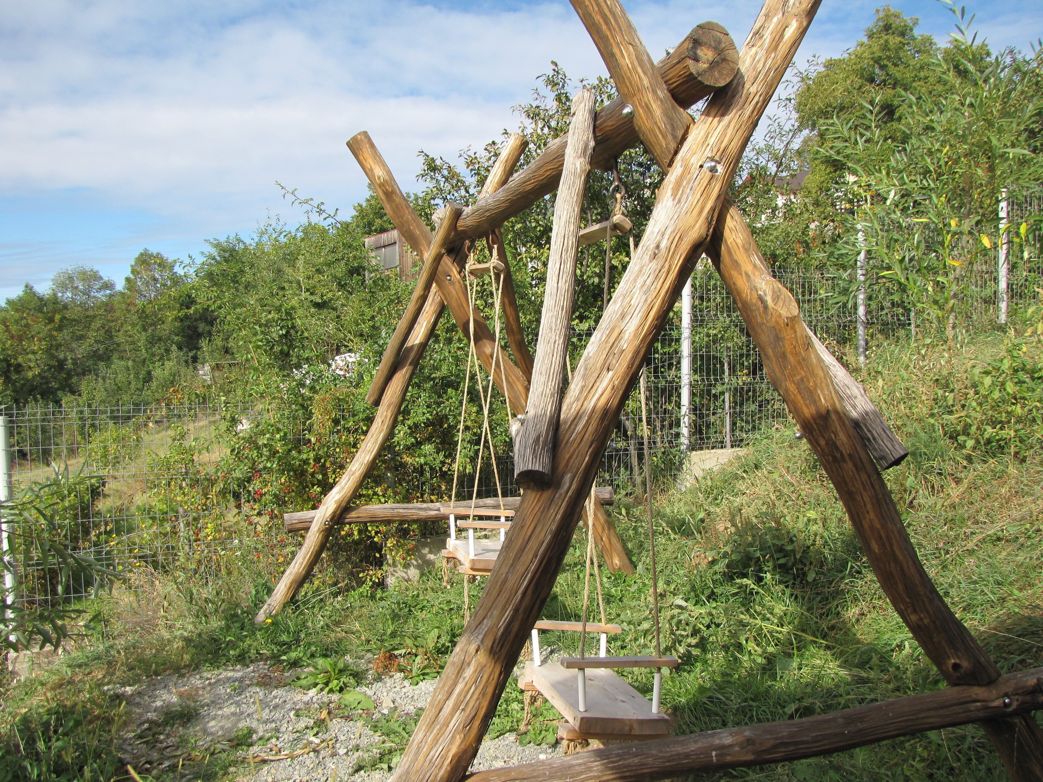 A wooden structure with a swing set, surrounded by greenery and a fence.