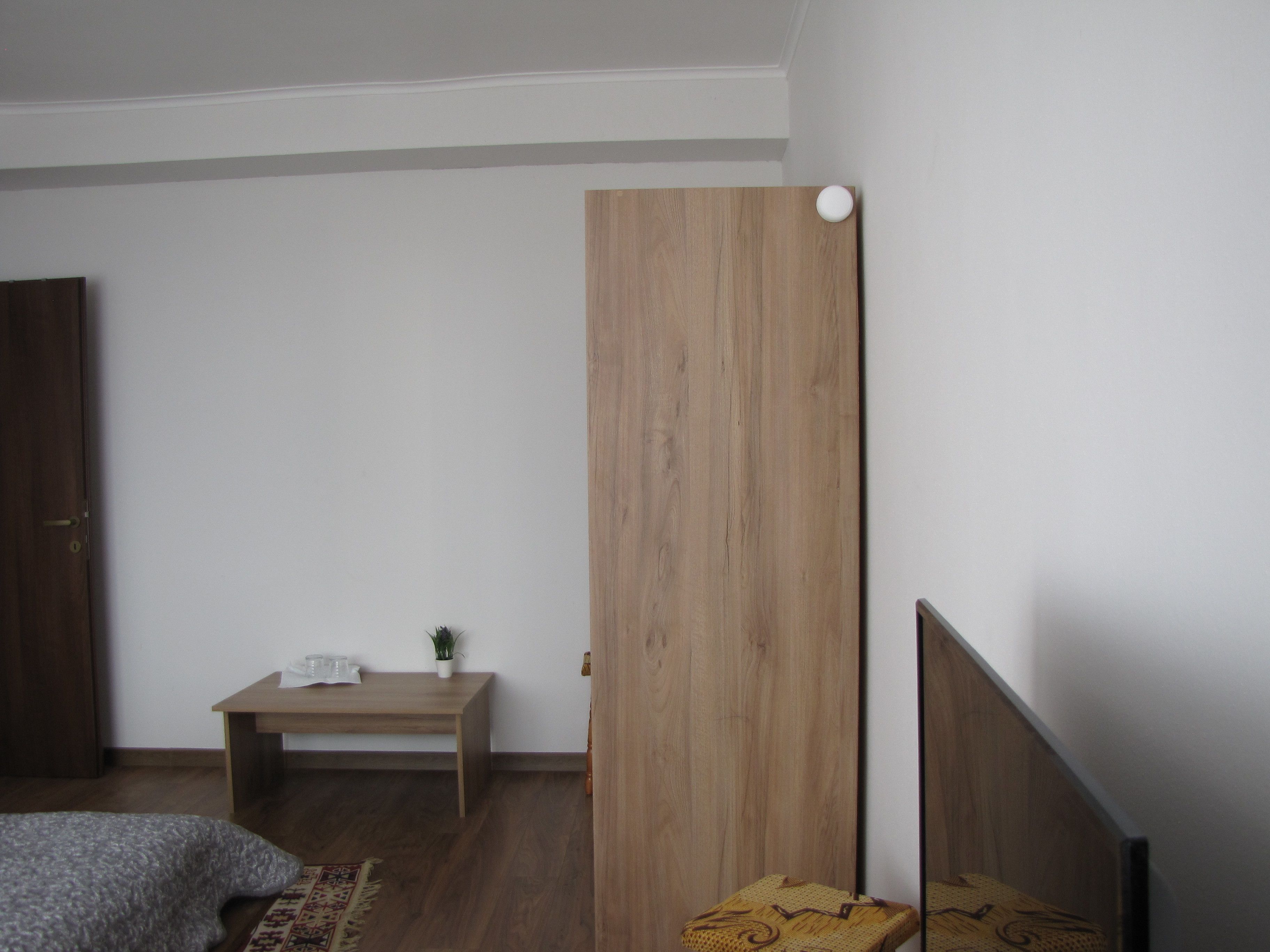 A bedroom with a wooden headboard, a bedside table with a plant, a bed with a gray blanket, and a wooden floor.
