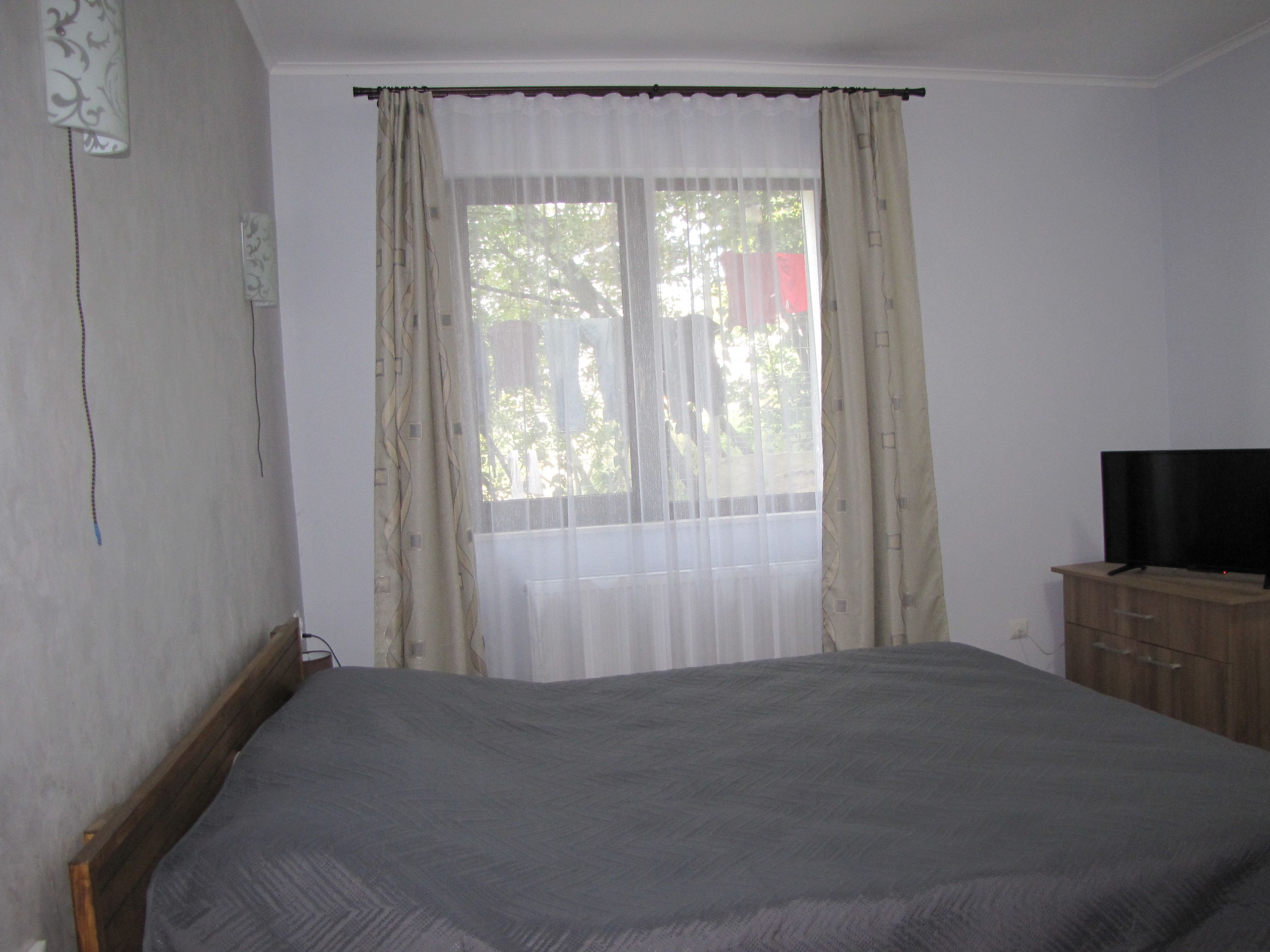 A bedroom with a bed, a television on a wooden cabinet, and a window with sheer curtains.