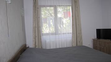 A bedroom with a bed, a television on a wooden cabinet, and a window with sheer curtains.