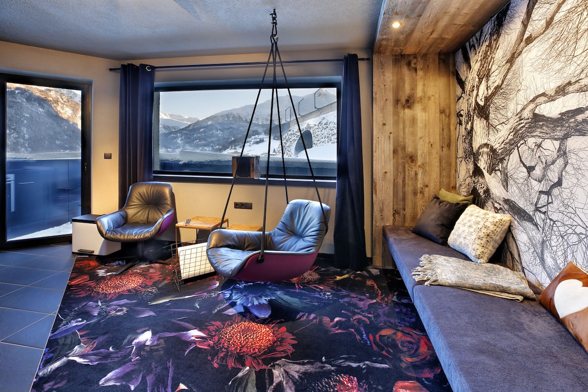 A modern interior with a sofa, a hanging chair, and a view of mountains through large windows.