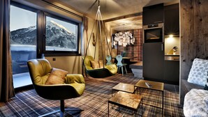 Un salon moderne doté d’une grande fenêtre, d’un canapé, d’un fauteuil, d’un luminaire suspendu et d’un coin cuisine.