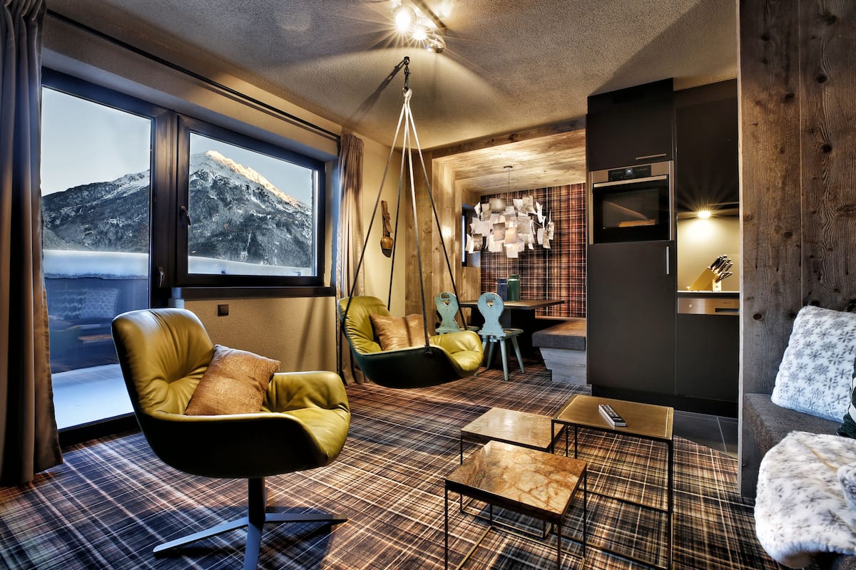 A modern living room with a large window, a sofa, a chair, a hanging light fixture, and a kitchen area.