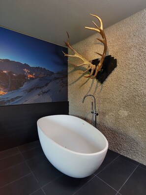 Ein modernes Badezimmer mit freistehender Badewanne, einem angebrachten Hirschgeweih und einem großen gerahmten Bild von einer Berglandschaft.
