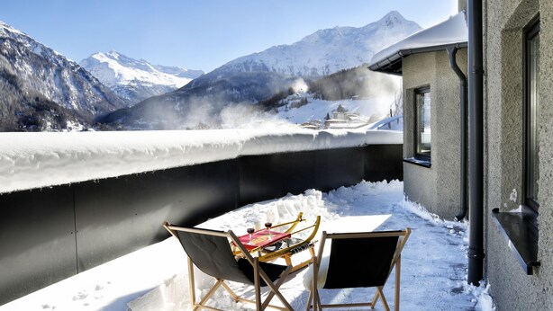 Un balcon enneigé avec vue sur les montagnes, deux chaises pliantes et une table recouverte d’une nappe rouge, ainsi que des verres à vin.