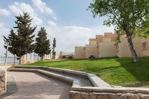 A paved pathway leading to a building with a stone wall and a grassy area with picnic tables. - Hi Mitzpe Ramon Hostel (Ramat Negev)