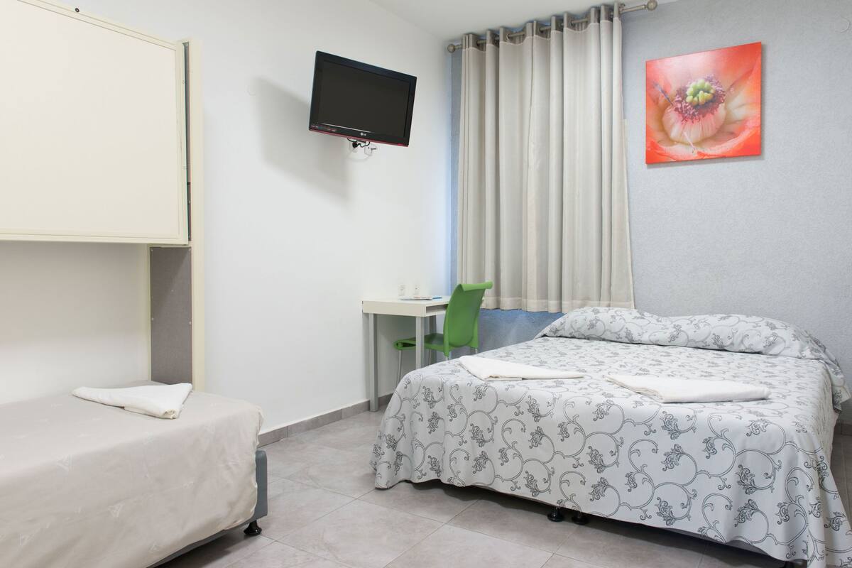 A hotel room with a single bed, a desk with a chair, a television mounted on the wall, and a window with curtains.