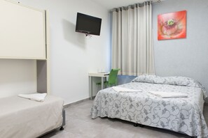 Une chambre d’hôtel avec un lit simple, un bureau avec une chaise, un téléviseur fixé au mur et une fenêtre avec des rideaux.