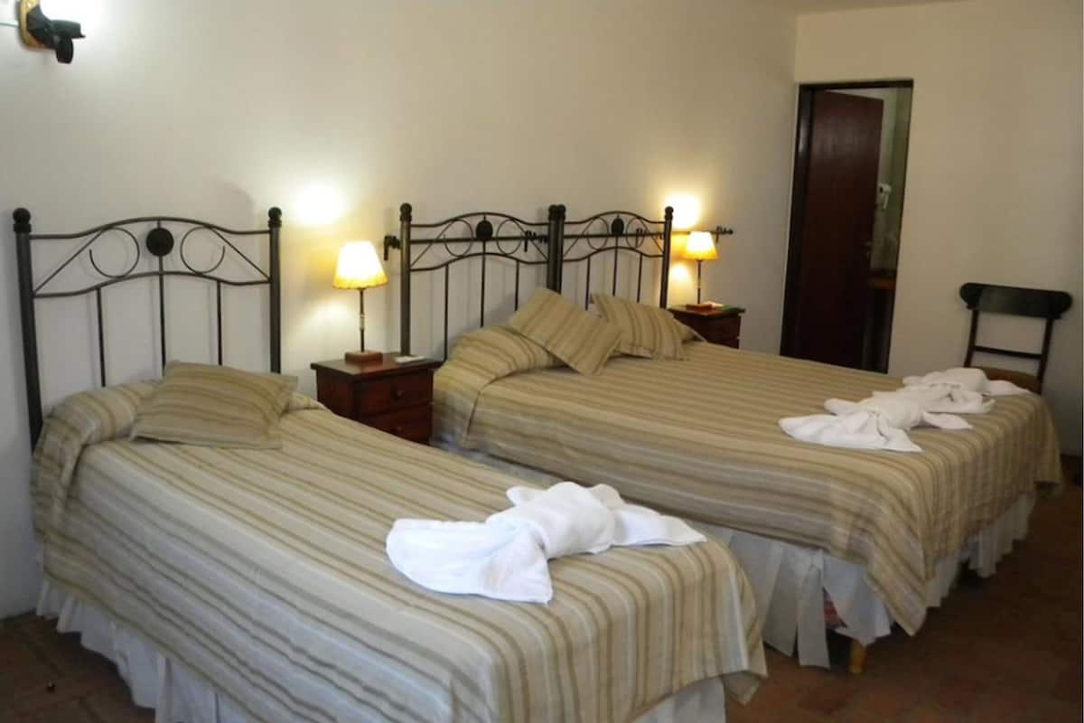 Quarto com duas camas, cada uma com colcha e almofadas listradas. Há mesas de cabeceira com abajures.