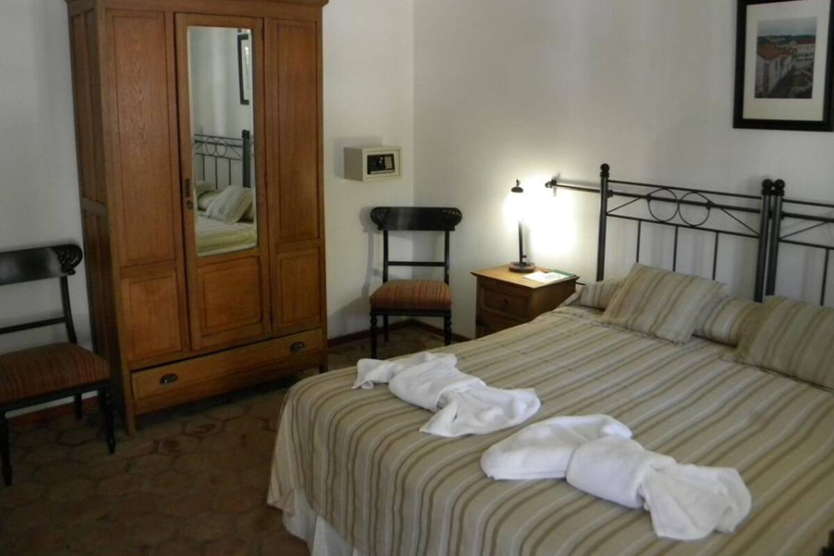 Um quarto com uma cama, um guarda-roupa, uma cadeira, uma mesa de cabeceira com abajur e um quadro pendurado na parede.