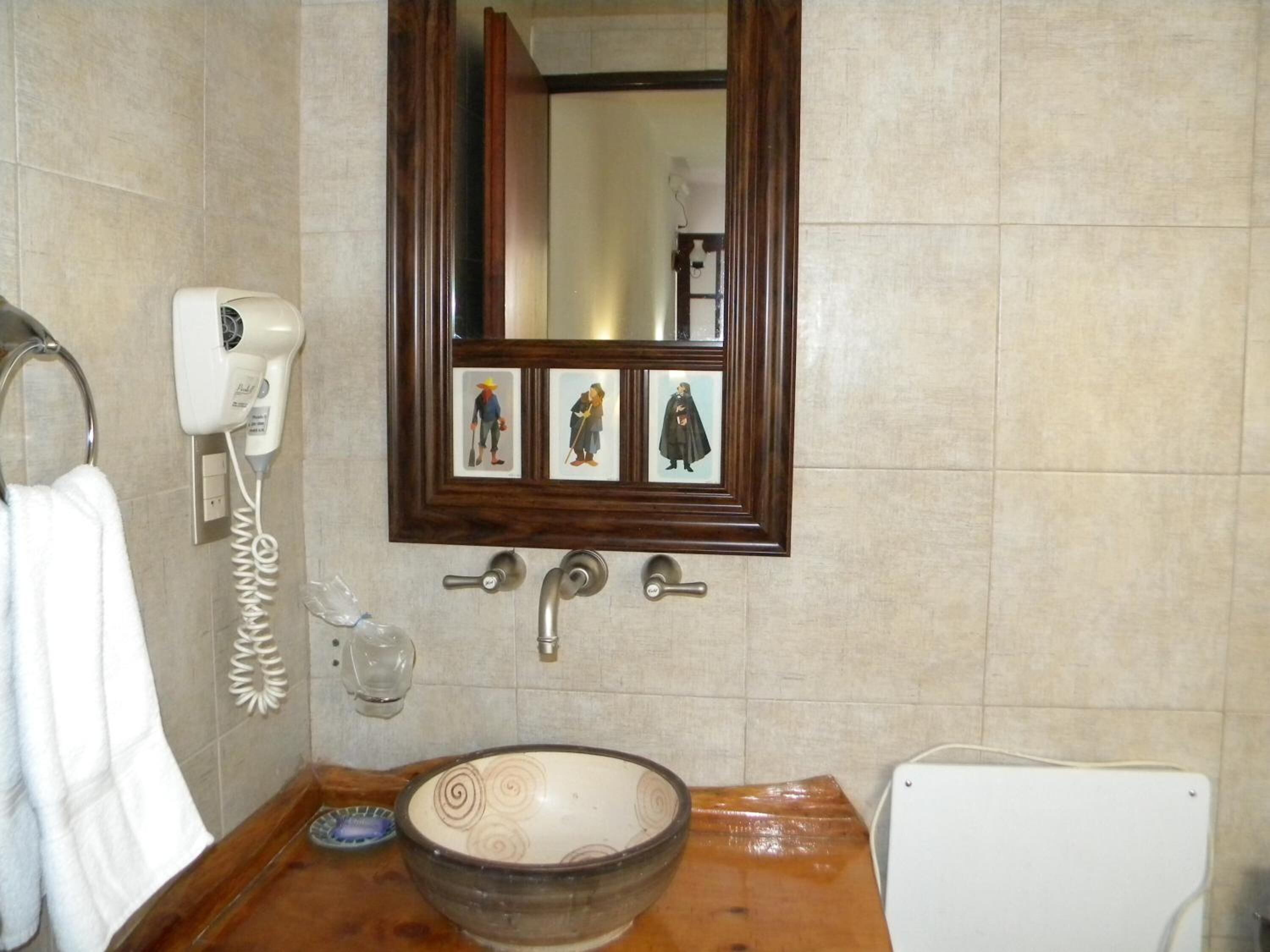 Un baño con tocador de madera, lavamanos blanco, espejo con fotografías enmarcadas y secador de pelo en la pared.