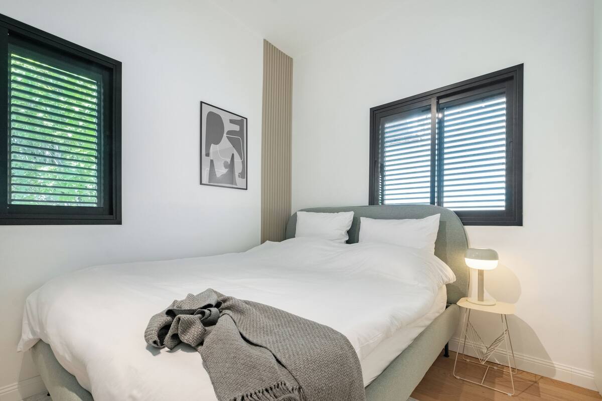 Ein Schlafzimmer mit einem Bett, einer Nachttischlampe und zwei Fenstern mit Jalousien.