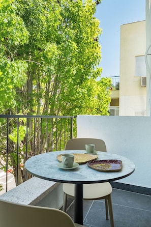A round table with two chairs, a cup, and a plate on a balcony with a view of trees and a building. - Mr. Yavos - By TLV2GO (Tel Aviv)