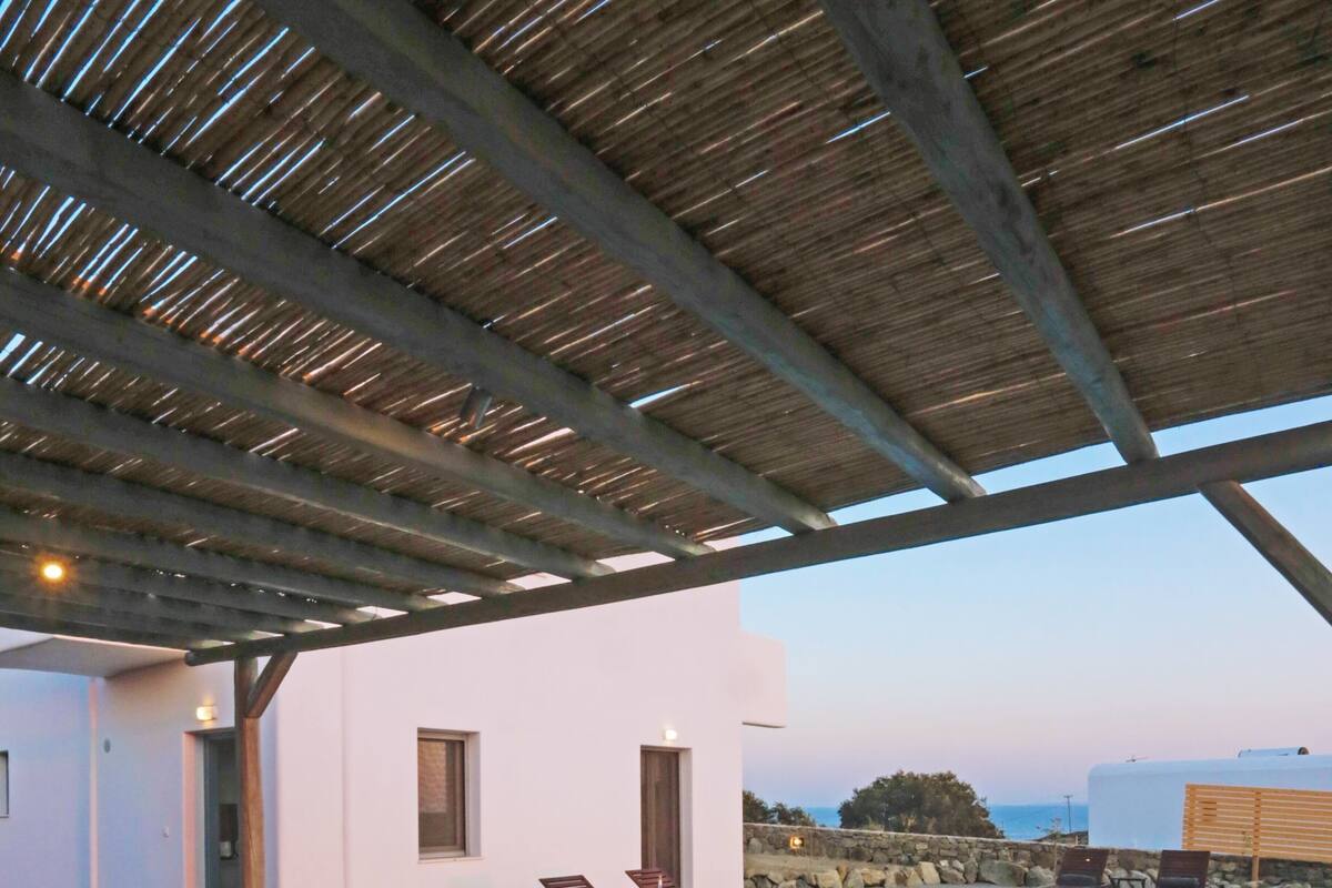 A covered outdoor area with a pool, wooden furniture, and a view of the sea.