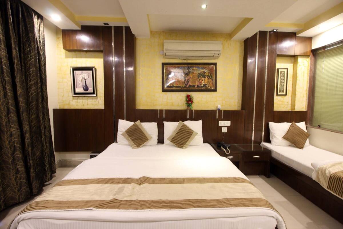 A hotel room with a large bed, two smaller beds, a nightstand, an air conditioning unit, and a framed picture on the wall.