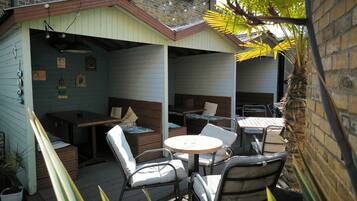 A covered outdoor seating area with wooden furniture and a table, surrounded by a brick wall and a wooden deck.