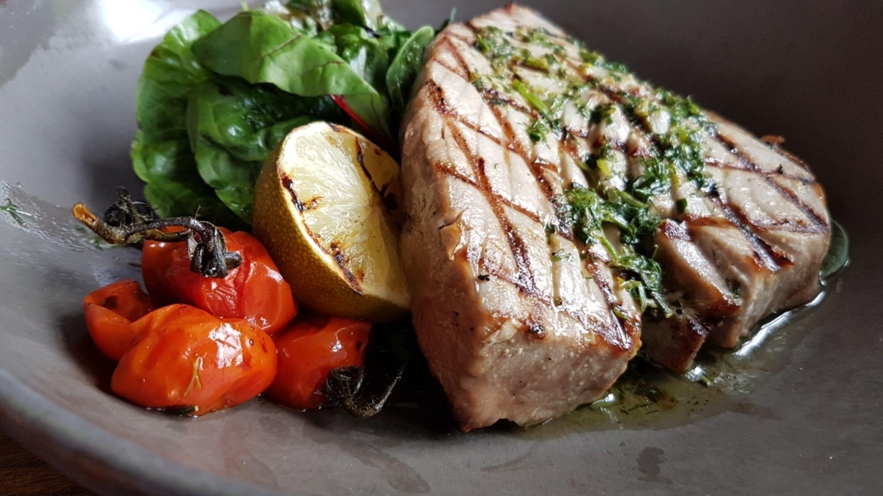 Un plato con pescado a la parrilla, tomates cherry, un limón y una ensalada.