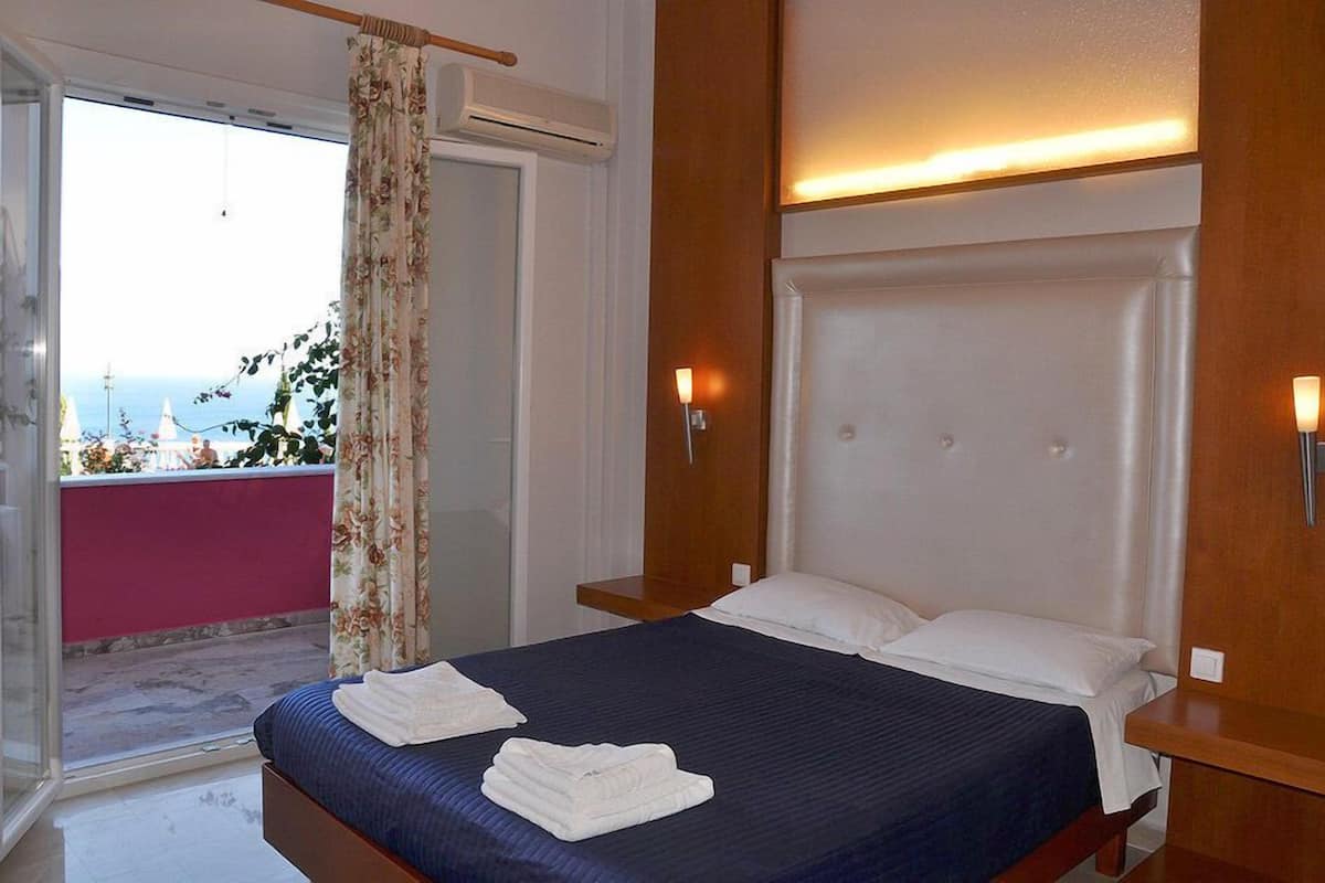 A hotel room with a bed, a wooden headboard, a nightstand, a window with curtains, and a view of the sea.