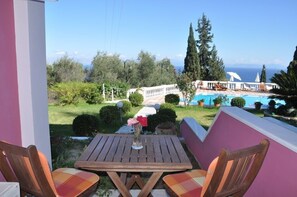 A patio with a wooden table and chairs, overlooking a pool and garden with a clear view of the sea. - Niki Apartments (Corfu)