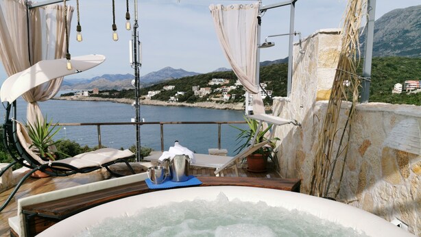 A hot tub with a view of the sea and mountains, a lounge chair, and a table with a bucket and pitcher.