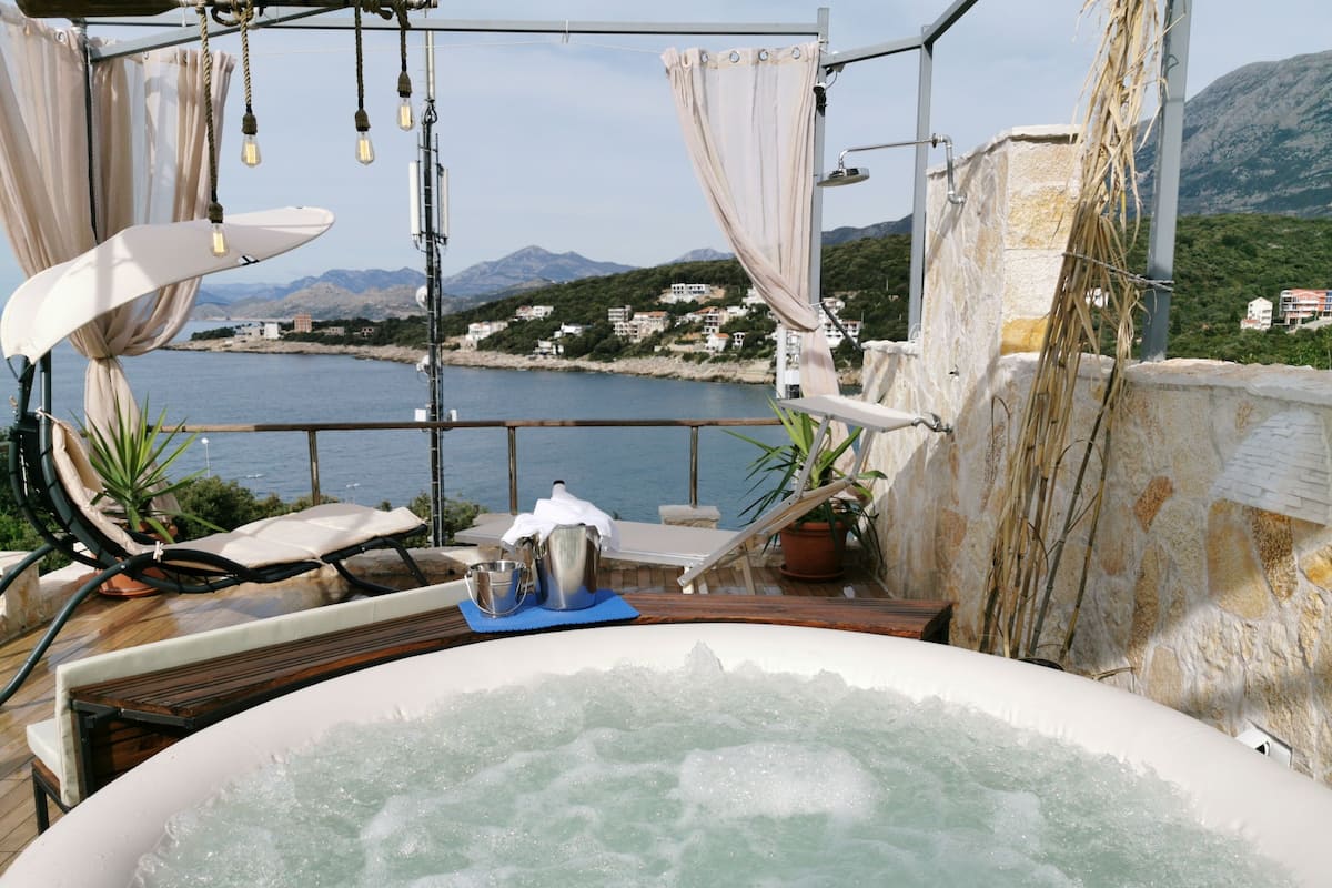 A hot tub with a view of the sea and mountains, a lounge chair, and a table with a bucket and pitcher.