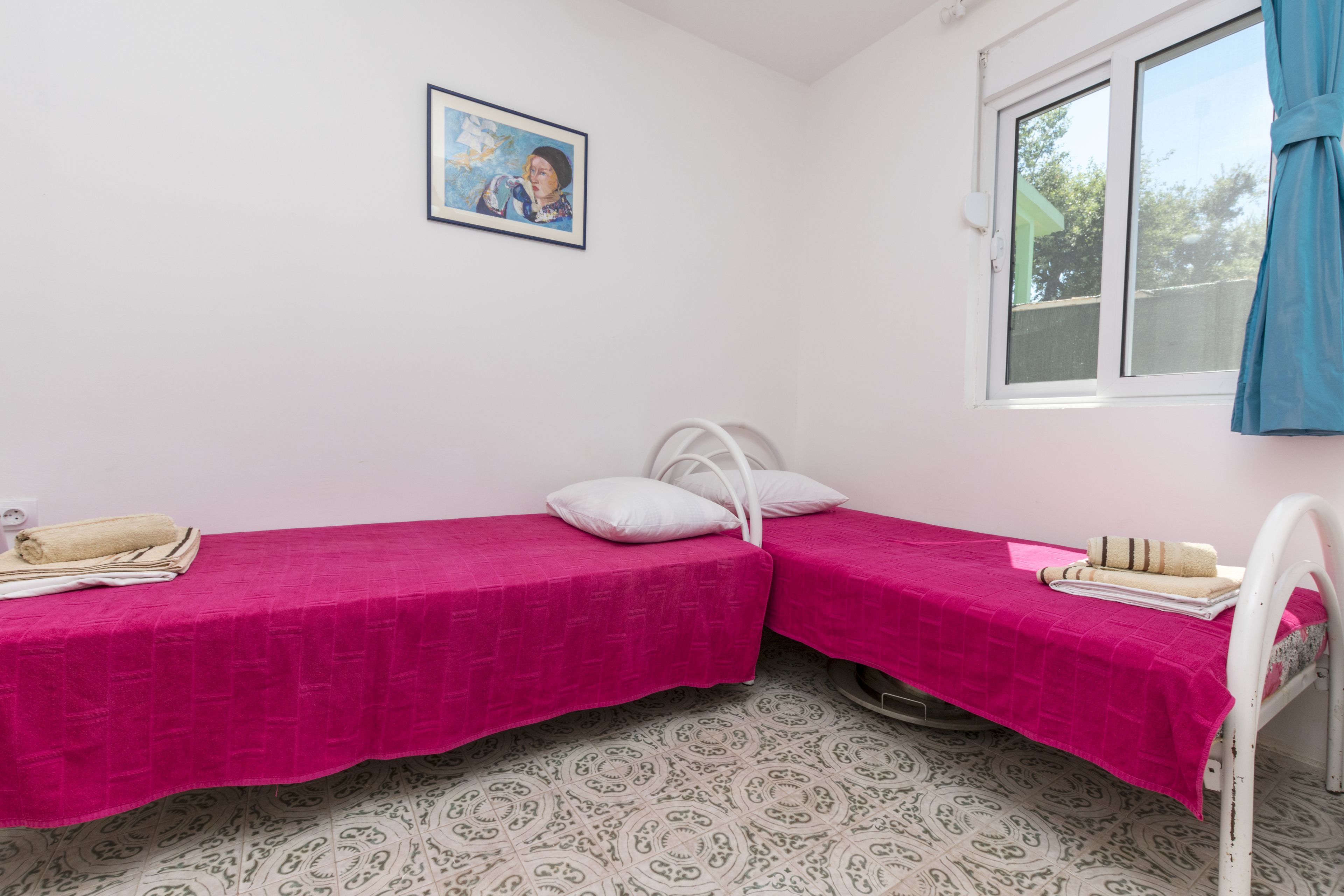 A small room with two beds, a window with blue curtains, a patterned floor, and a framed picture on the wall.