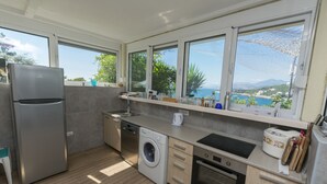 A modern kitchen with a built-in washing machine, dishwasher, and oven. It features large windows offering a view of the sea and mountains.