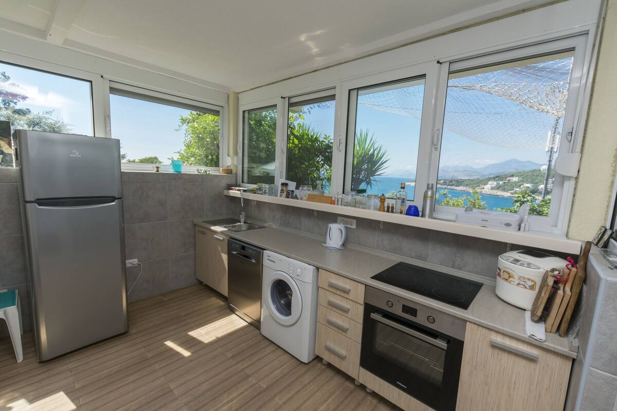 A modern kitchen with a built-in washing machine, dishwasher, and oven. It features large windows offering a view of the sea and mountains.