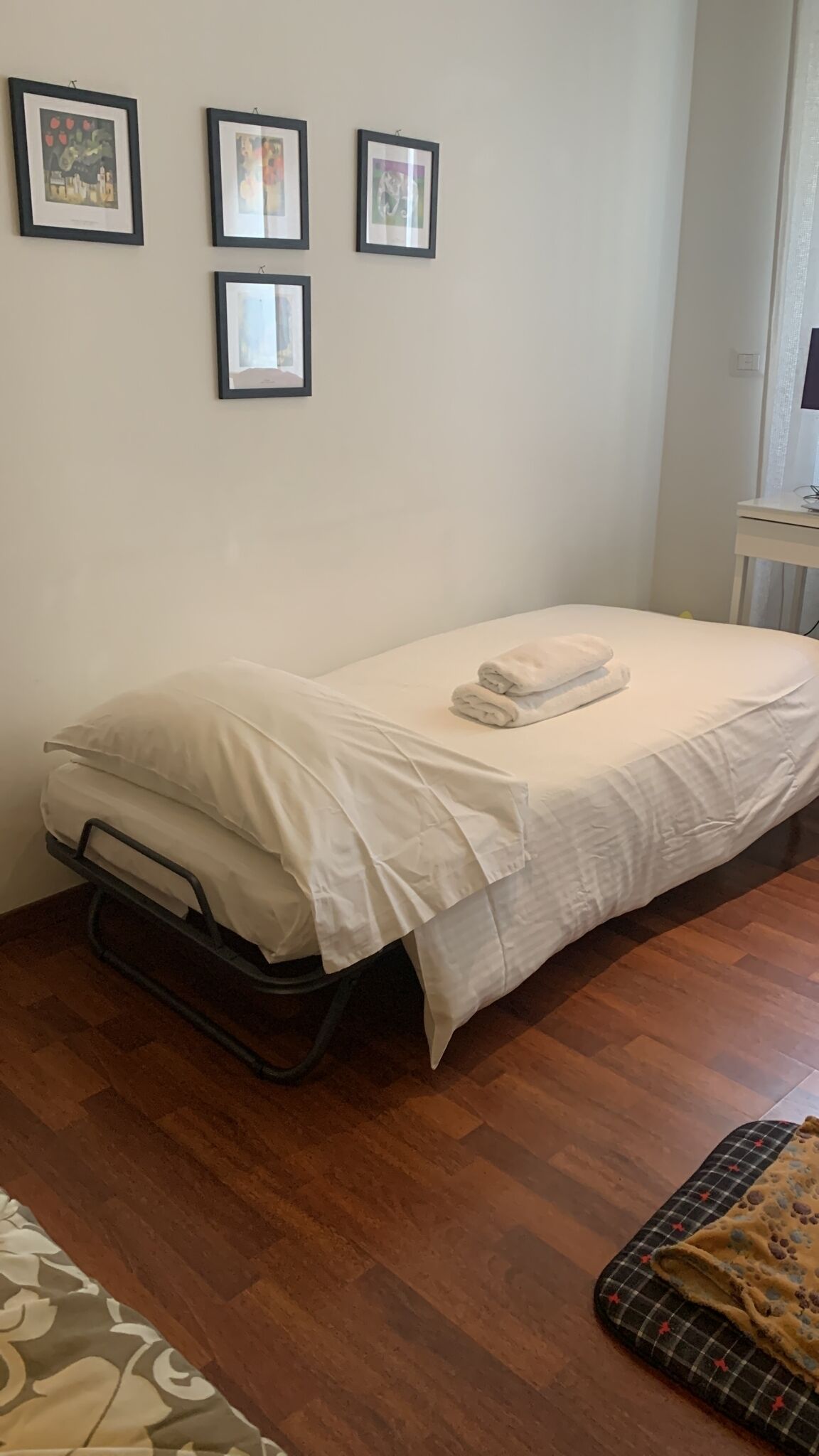 A small, neatly made bed with white bedding and a folded towel on top. There are three framed pictures on the wall above the bed.