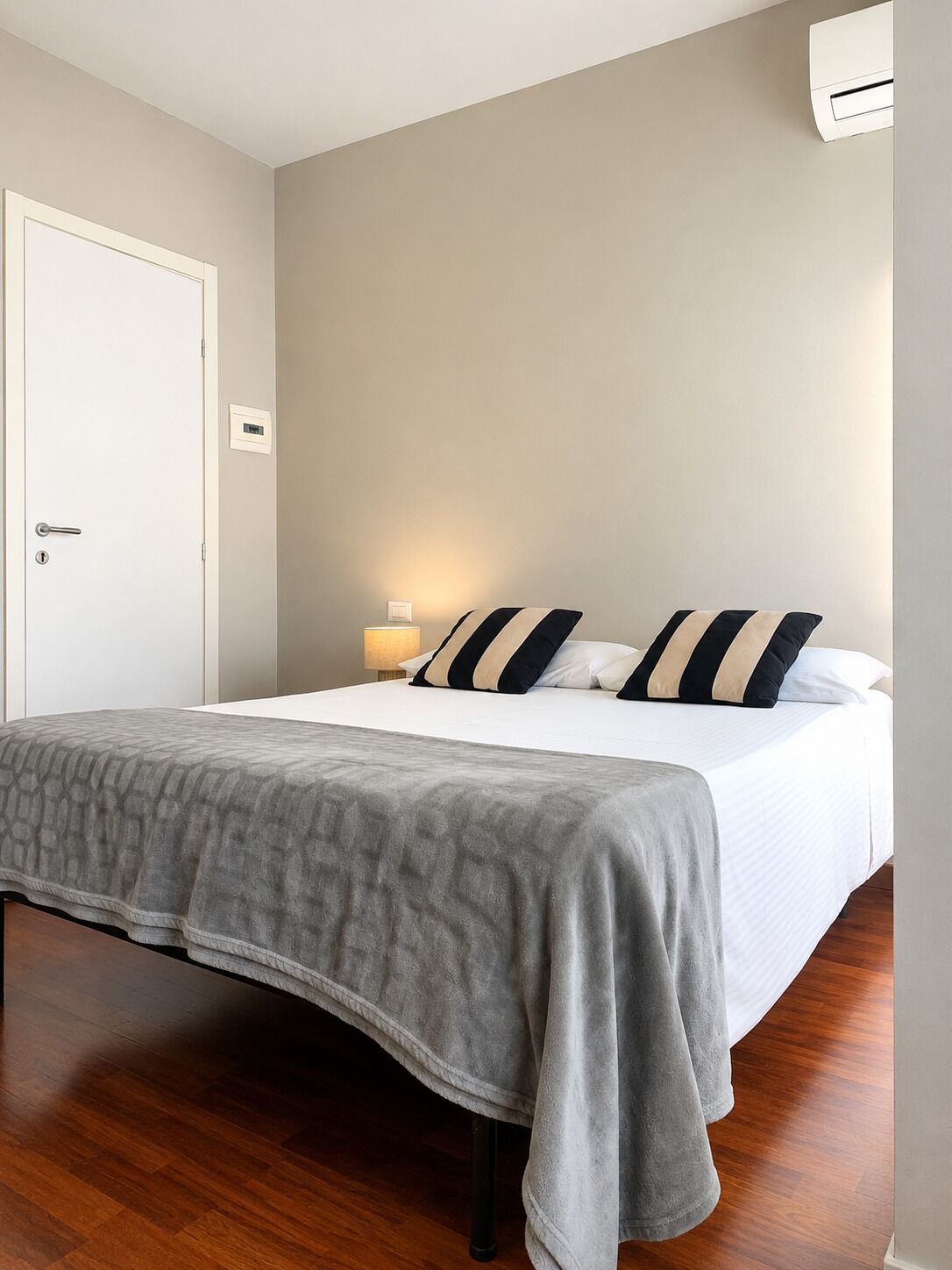 A single bed with a grey blanket, two pillows with black and white stripes, a bedside lamp, and a white air conditioning unit on the wall.