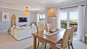 Ein modernes Wohnzimmer mit Esstisch aus Holz, Korbstühlen, einer Couch und einem an der Wand befestigten Fernseher.