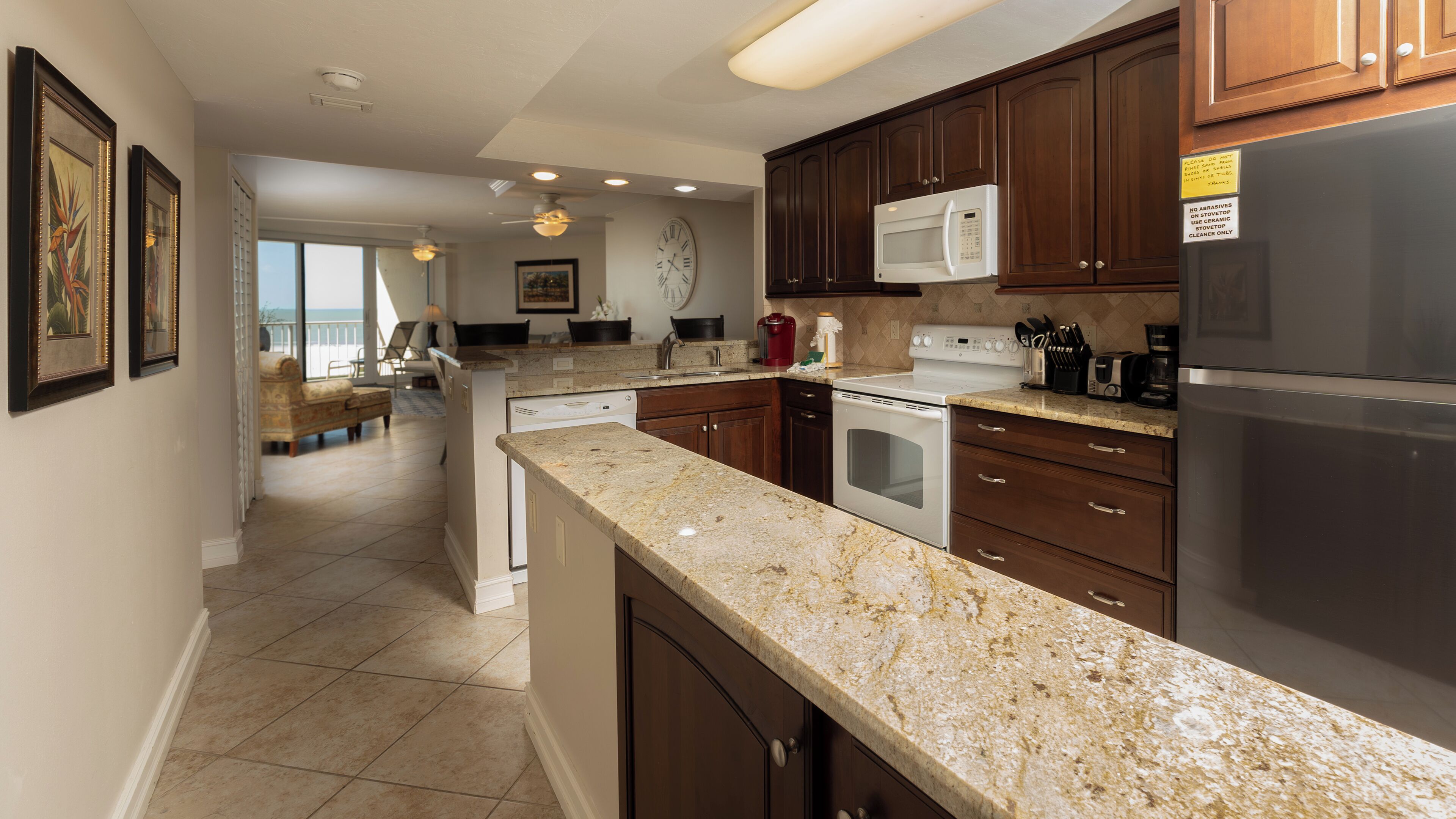 A kitchen with dark wooden cabinets, a white refrigerator, a microwave, and a stove. There is a dining area with a view of the ocean.