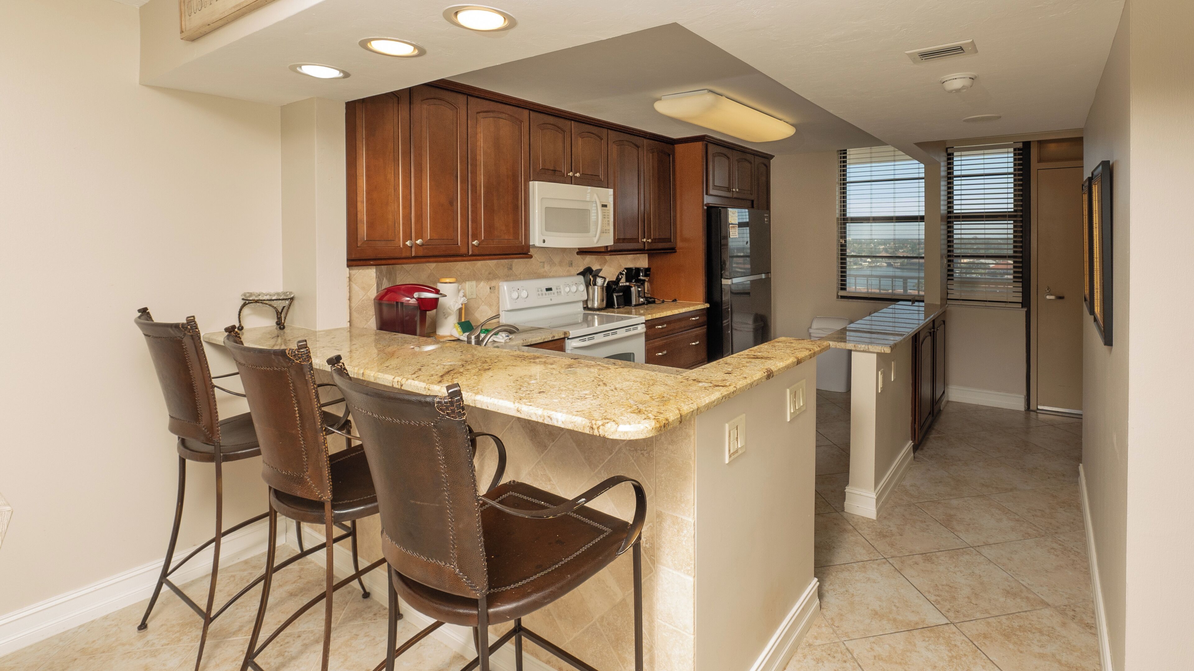A kitchen with wooden cabinets, a white microwave, a stove, and a refrigerator. There are three bar stools and a window with blinds.