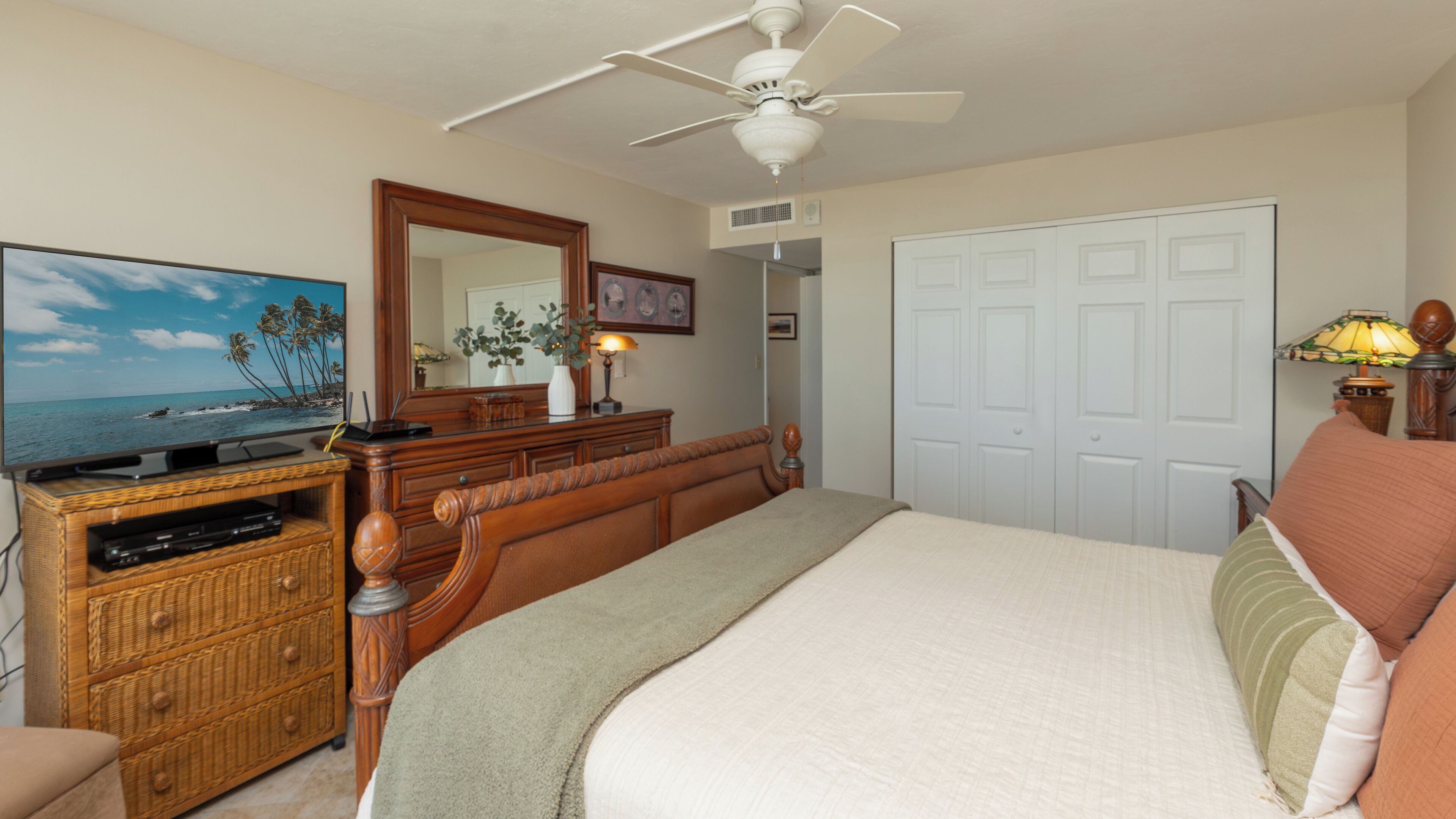 Une chambre à coucher comprenant un lit, une commode, un téléviseur, un ventilateur de plafond et une porte.