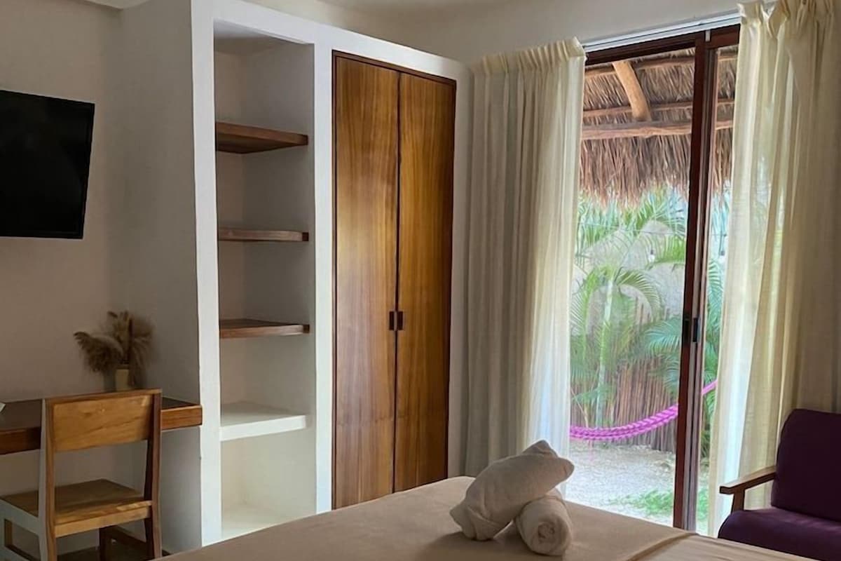 A hotel room with a bed, a desk, a chair, a wardrobe, and a sliding glass door to a balcony.