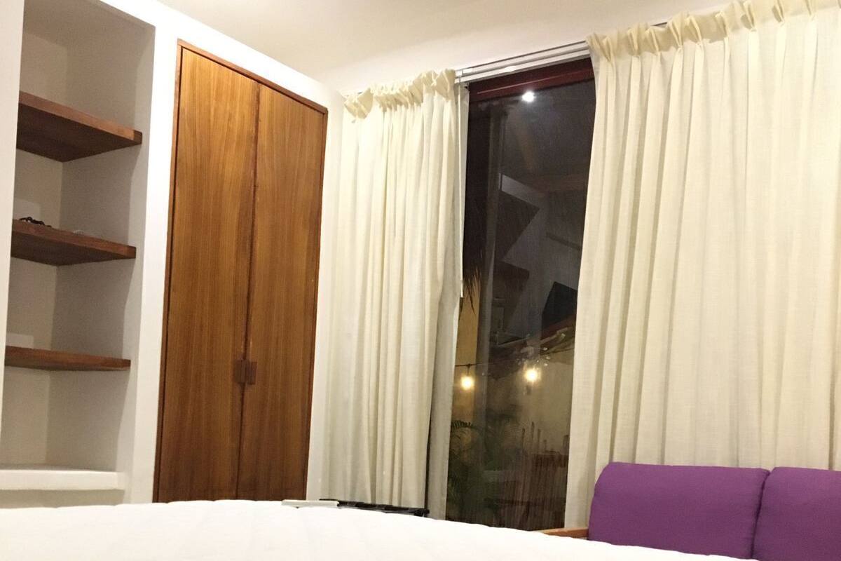 Un dormitorio con cama, armario, sofá morado y ventana con cortinas.
