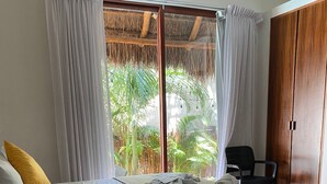 A room with two beds, a wooden wardrobe, and a view of a tropical garden through a large window with curtains. - Hotel Pelecanus Suites (Isla Holbox)