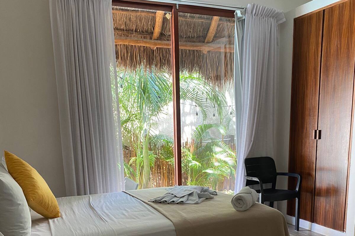 Una habitación con dos camas, un armario de madera y vista a un jardín tropical a través de un ventanal con cortinas.