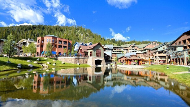 Un hotel frente al lago con un puente de piedra, edificios que se reflejan y montañas boscosas al fondo.