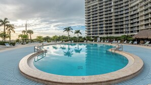 Ein großer, kreisförmiger Swimmingpool mit einem Sprungverbotsschild, umgeben von Liegestühlen und Palmen, mit einem Hochhaus im Hintergrund.
