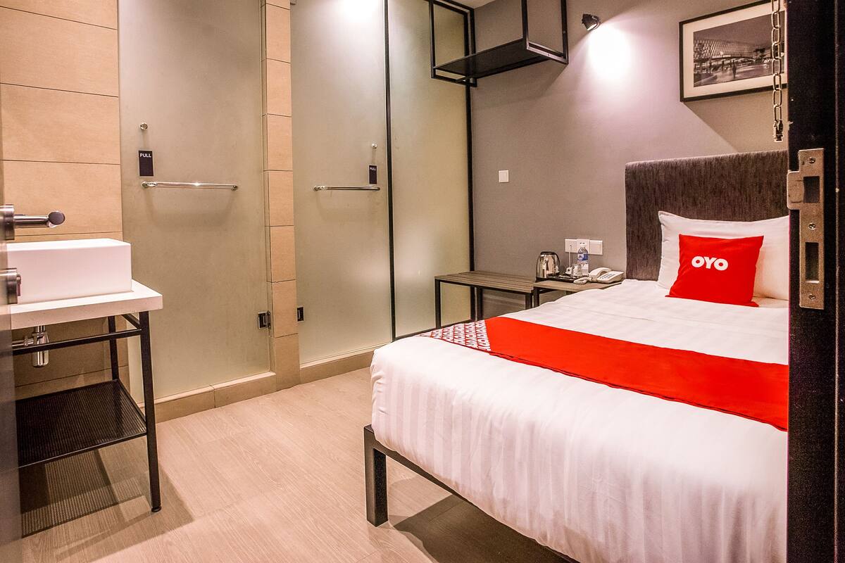 Une chambre d’hôtel comprenant un lit, une table de chevet avec un téléphone et une bouilloire, une salle de bain avec un lavabo et un miroir, et une porte dotée d’une poignée.