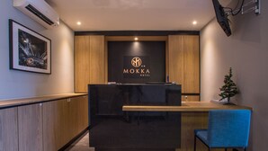 Un mostrador de recepción con un letrero que dice "MOKKA HOTEL" en el centro, paneles de madera en las paredes, un pequeño árbol de Navidad y una foto enmarcada a la izquierda.
