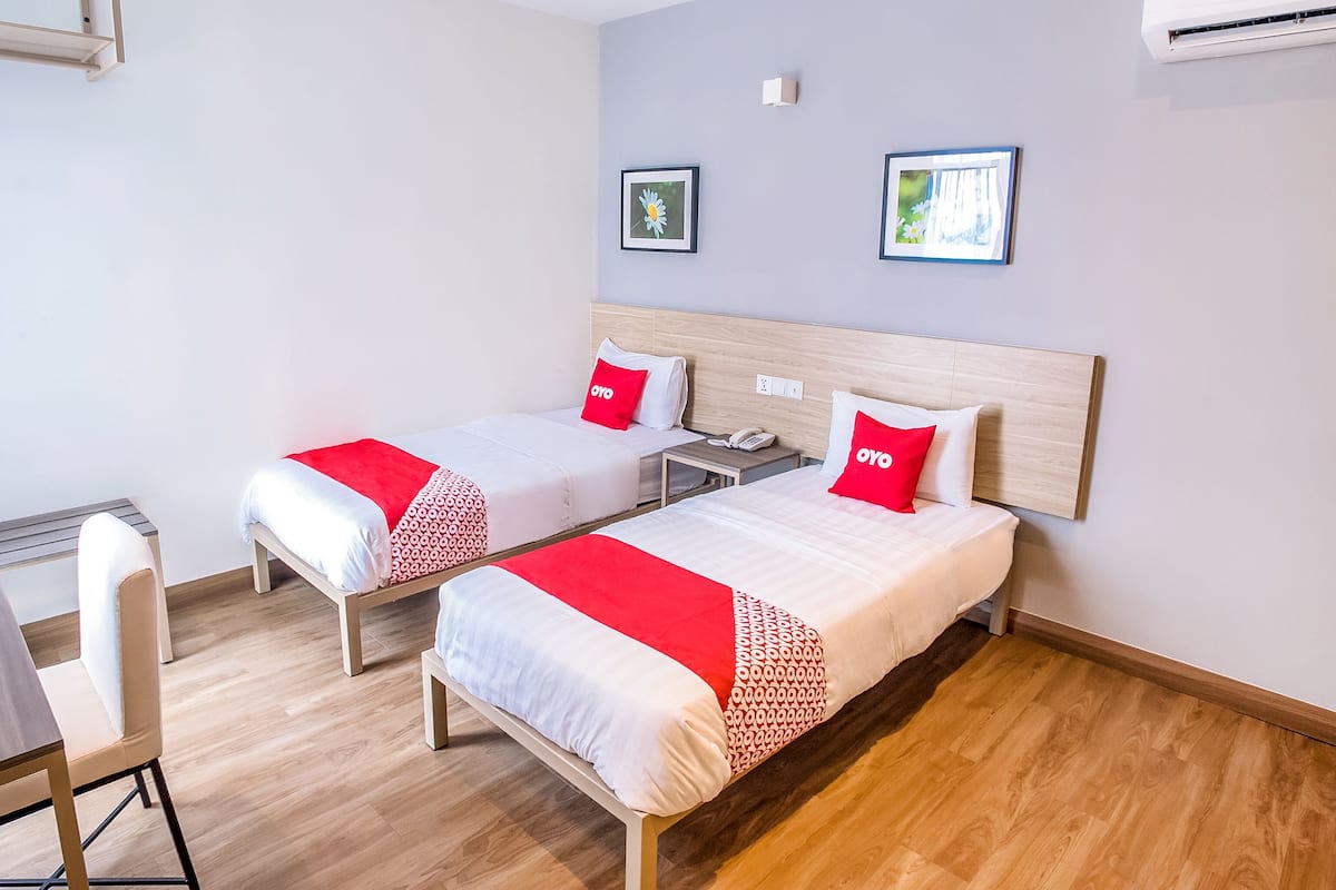 Une chambre d’hôtel avec deux lits, chacun recouvert d’une couvre-lit à motifs rouges et blancs, assortie aux oreillers. Il y a une petite table de chevet avec un téléphone et une lampe. Les murs de la chambre sont de couleur claire et le sol est en bois.